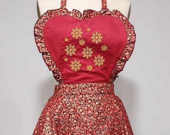 May include: A red and gold Christmas apron with a heart-shaped top and a snowflake design. The apron has a ruffled trim and a tie at the waist.