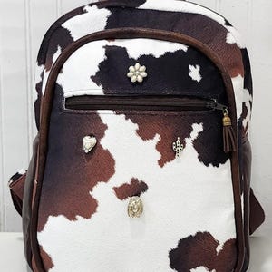 May include: A brown and white cow print backpack with a brown leather trim. The backpack has a zippered pocket on the front and several decorative metal accents.