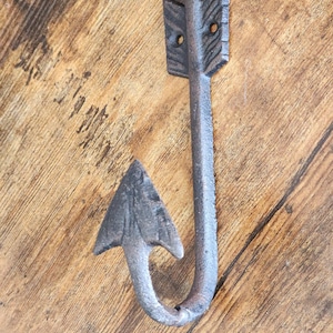 May include: A cast iron arrow hook with a rustic finish. The hook is shaped like an arrow with a pointed tip and a curved hook at the bottom. The arrow is decorated with a simple design.