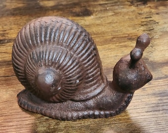 May include: A cast iron snail figurine in a rustic brown finish. The snail has a detailed shell with textured ridges and prominent eye stalks. The figurine is designed to sit on a flat surface.