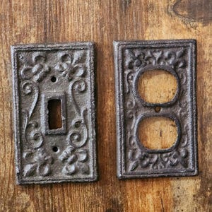 May include: Two cast iron switch plates with an ornate floral design. One plate has a single switch opening, and the other has two outlet openings. The plates are dark gray and sit on a wooden surface.