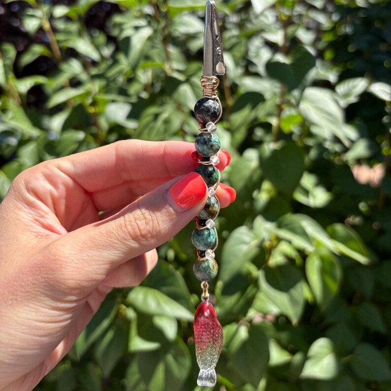 May include: A metal alligator clip roach clip featuring a vertical arrangement of round, mottled green and black stone beads separated by small clear glass spacers, finished with a translucent red and clear glass fish charm at the bottom.