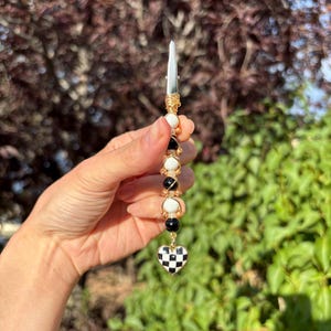 May include: A metal roach clip featuring a vertical arrangement of alternating black and white spherical beads wrapped in gold-toned wire. The bottom of the accessory terminates in a heart-shaped charm with a black and white checkered pattern.