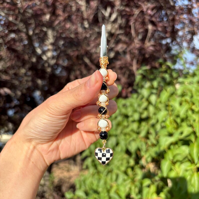 May include: A metal roach clip featuring a vertical arrangement of alternating black and white spherical beads wrapped in gold-toned wire. The bottom of the accessory terminates in a heart-shaped charm with a black and white checkered pattern.