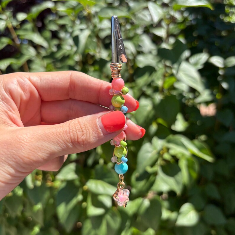 May include: A metal alligator clip attached to a decorative beaded stem featuring a variety of round beads in shades of pink, lime green, and light blue, finished with a small, clear, faceted star-shaped charm dangling at the bottom end.