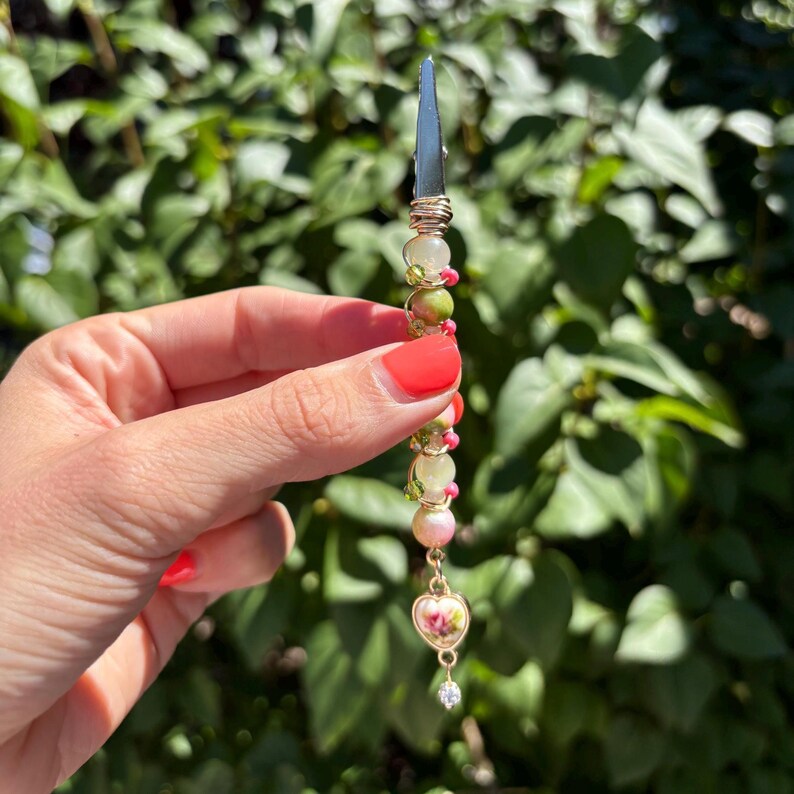 May include: A metal roach clip featuring a vertical arrangement of green, pink, and cream-colored beads wrapped in gold-toned wire. The accessory terminates in a small heart-shaped charm with a floral pattern and a tiny clear crystal dangle.