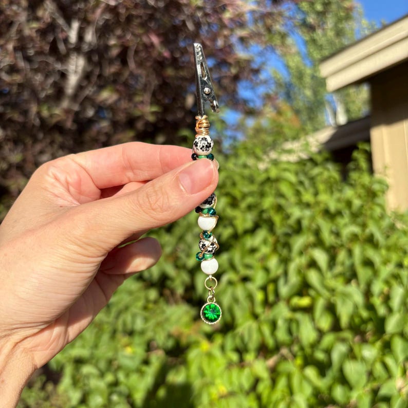 May include: A metal roach clip featuring a vertical arrangement of beads in white, black, and green, accented with gold-toned wire wrapping. The accessory terminates in a circular, faceted emerald green charm at the bottom end of the beaded stem.