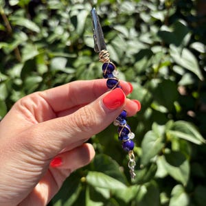 May include: A metal alligator clip adorned with a series of dark blue, light blue, and clear beads wrapped in gold-toned wire. The decorative clip features a small star-shaped charm dangling from the bottom end of the beaded wire structure.