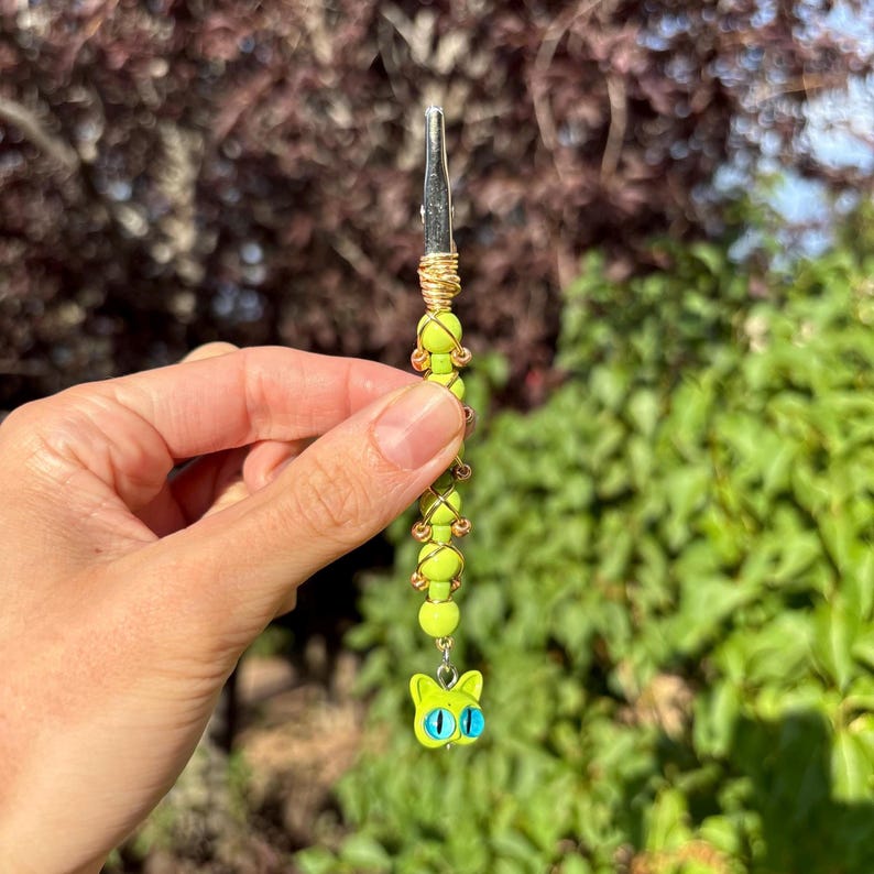 May include: A metal roach clip featuring a series of bright lime green beads wrapped in thin gold-toned wire. The bottom of the accessory is finished with a small, lime green cat-shaped charm that has two prominent, round, bright blue eyes.