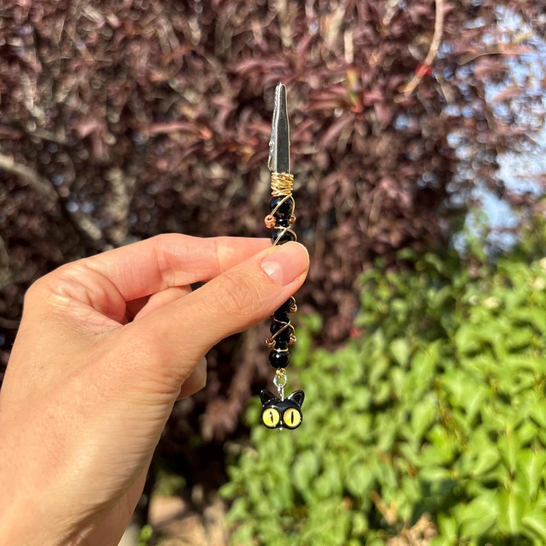 May include: A metal roach clip featuring a series of round black beads wrapped in thin gold-toned wire. The end of the clip is adorned with a small black cat charm that has prominent, bright yellow eyes, creating a distinct decorative accessory.