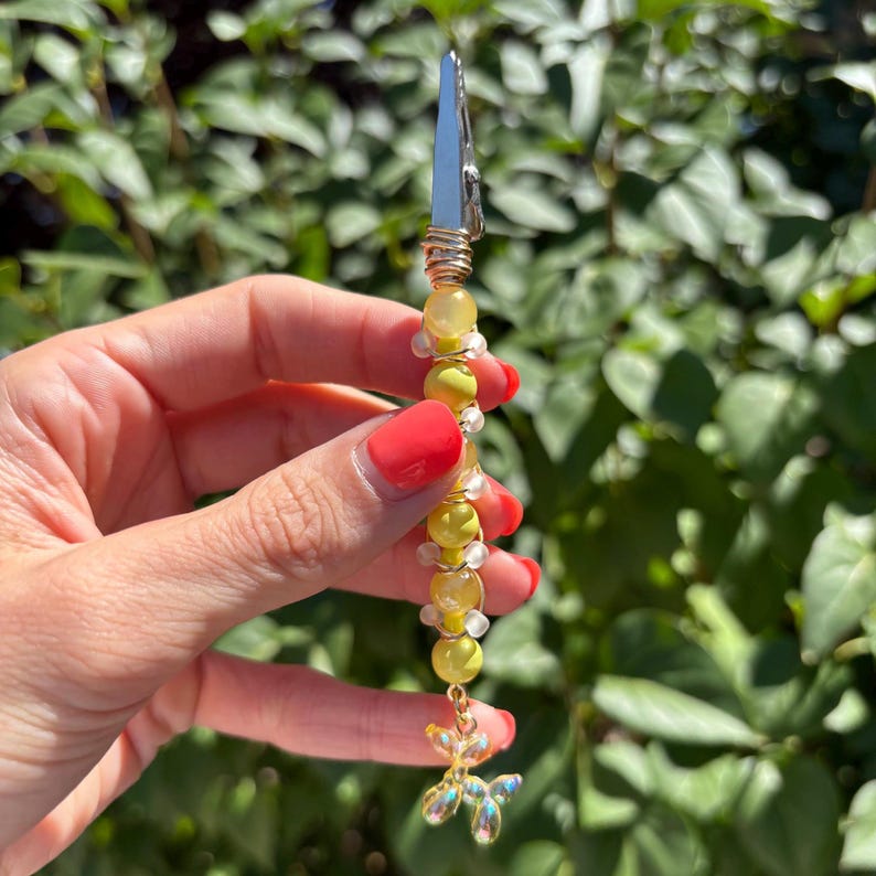 May include: A metal alligator clip roach clip featuring a vertical arrangement of yellow and translucent beads wrapped in gold-toned wire, finished with a small, iridescent, clear butterfly-shaped charm dangling at the bottom end.