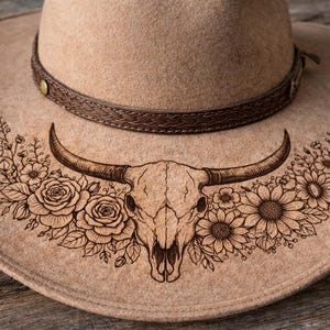 May include: A tan cowboy hat with a brown leather band. The hat features a detailed wood-burned design of a bull skull with floral accents, including roses and sunflowers.
