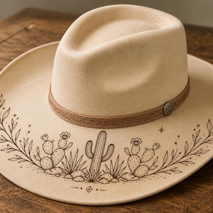 May include: A beige cowboy hat with a brown leather band and a decorative metal button. The hat features a detailed desert scene with cacti and foliage in a dark brown color, adding a touch of Southwestern style.