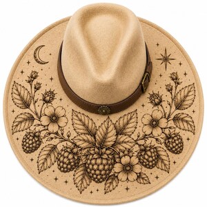 Könnte beinhalten: Beigefarbener Fedora-Hut mit braunem Lederband und Schnalle. Der Rand ist mit einem geätzten Design aus Himbeeren, Blumen, Blättern, einem Mond und einem Stern verziert. Ein modischer Hut.