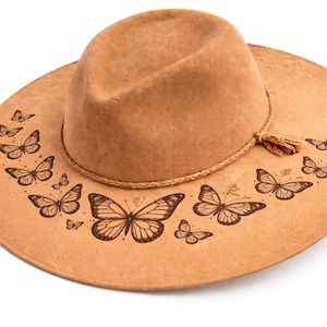 May include: A tan-coloured fedora hat with a wide brim. The brim is decorated with a pattern of detailed butterfly illustrations in a darker brown colour. The hat has a rope band with a tassel.