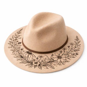 Peut inclure: Un chapeau fedora beige avec un bandeau marron et un motif floral. Le chapeau a un large bord avec un motif floral noir sur le bord. La couronne est de couleur beige uni.