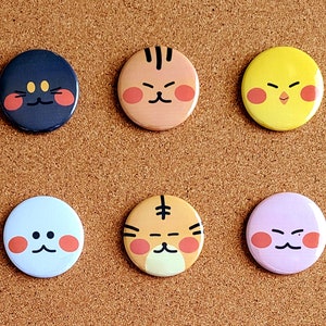 Könnte beinhalten: Sechs runde Anstecknadeln mit verschiedenen Comic-Gesichtern. Die Gesichter sind gelb, orange, weiß, lila, schwarz und hellbraun. Jedes Gesicht hat rote Wangen und einen anderen Ausdruck.