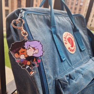 Puede incluir: Un llavero acrílico transparente con una ilustración de dibujos animados de dos personajes abrazándose, con corazones rosas. El llavero está sujeto a un cierre en forma de corazón de oro rosa. Un mochila azul en el fondo.