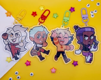 May include: Four colorful cartoon character keychains featuring characters from the animated series The Owl House. The characters are:  Hunter,  Eda,  Luz, and  Amity.  Each keychain is made of clear acrylic and has a metal ring for attaching to a bag or backpack.