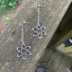May include: A pair of silver dangle earrings with atom-shaped charms. The charms have a silver outline and are decorated with colorful rhinestones.