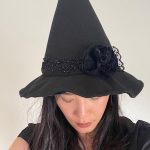 May include: A black witch hat with a wide brim and a tall, pointed crown. The hat is decorated with a black lace band and a large, dark rose. The hat is a costume accessory.