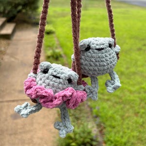 May include: Two light gray crocheted frog figures hanging from brown yarn ropes. One frog wears a ruffled pink skirt around its waist. Both frogs feature small black bead eyes and simple stitched mouths, crafted from soft yarn in a plush style.