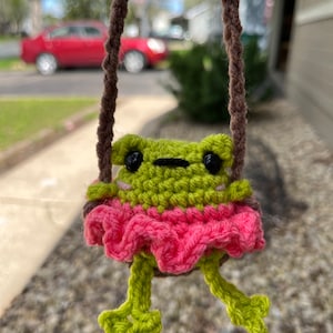 May include: A handmade crochet frog ornament in a swing. The frog is green with black eyes and a pink skirt. The swing is made of brown yarn. The background is blurred, showing a street and a red car.