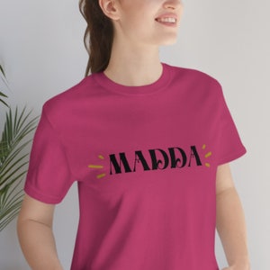 Peut inclure: Un t-shirt rose avec le mot "MADDA" imprimé en noir avec des accents jaunes.