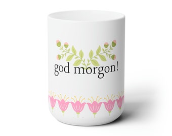 May include: White ceramic mug with the text "god morgon!" in black. The mug is decorated with a floral design featuring pink flowers and green leaves. A decorative and cheerful coffee mug.