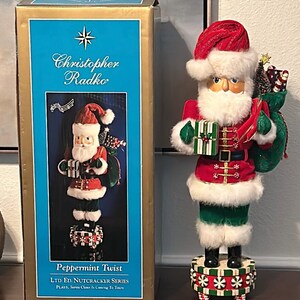 May include: A decorative Santa Claus figurine with a red and white hat, red coat, and green pants. The figurine holds a green gift box and a green sack. The box next to the figurine has the text "Christopher Radko" and "Peppermint Twist".