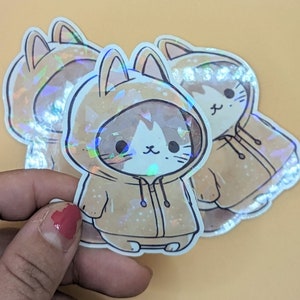 Könnte beinhalten: Holografische Aufkleber mit einer Cartoon-Katze, die einen beigefarbenen Kapuzenpullover trägt. Die Katze hat ein weißes und braunes Gesicht mit schwarzen Augen und Schnurrhaaren. Die Aufkleber haben ein glänzendes, irisierendes Finish und sind auf hellorangefarbenem Hintergrund.
