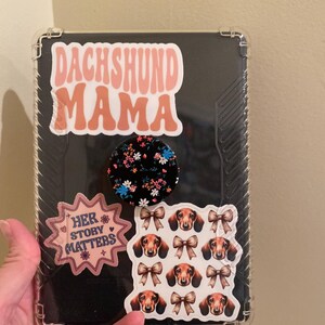 May include: A clear phone case with a pink and white "Dachshund Mama" sticker, a black and white floral sticker, a pink sticker that says "Her Story Matters", and a sticker with multiple images of dachshunds with bows.