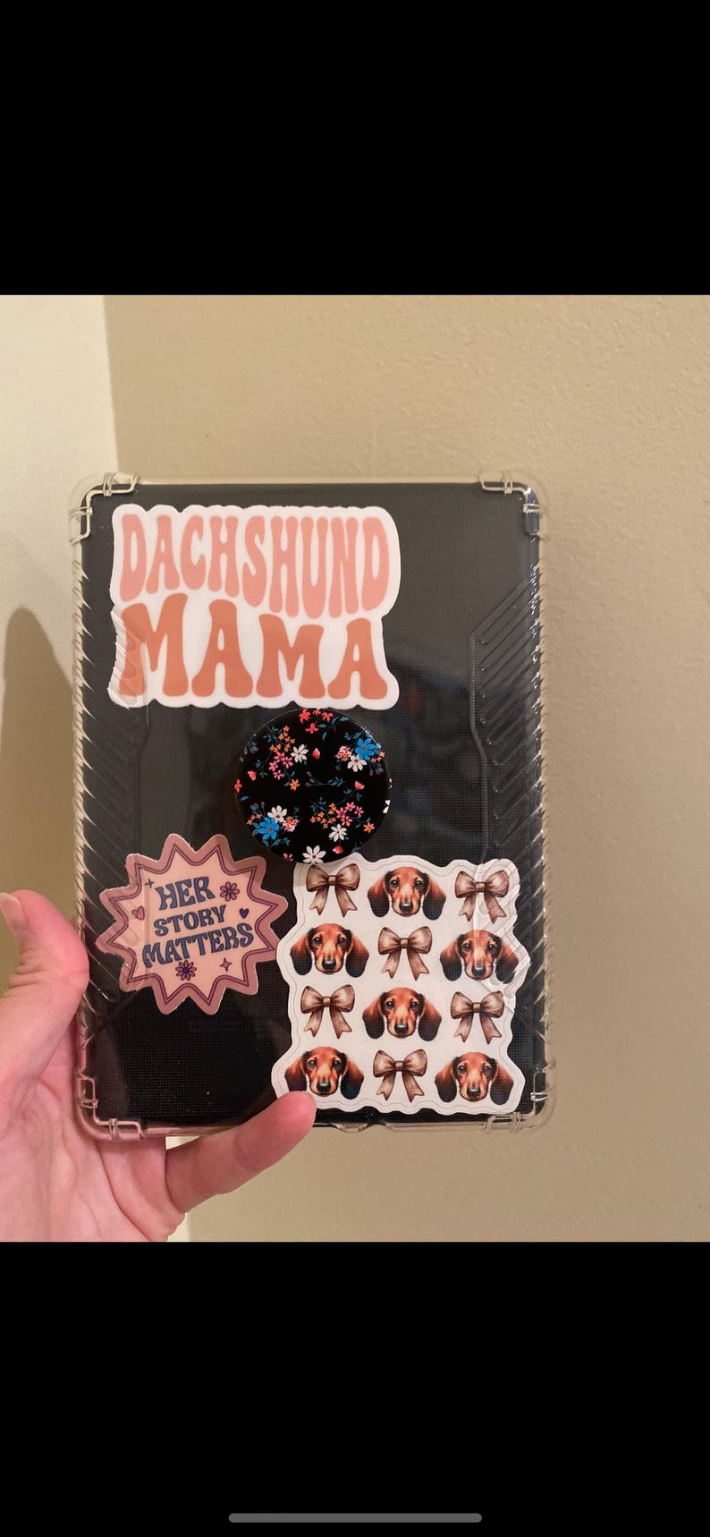 May include: A clear phone case with a pink and white "Dachshund Mama" sticker, a black and white floral sticker, a pink sticker that says "Her Story Matters", and a sticker with multiple images of dachshunds with bows.