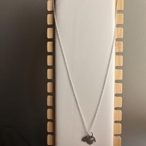 Puede incluir: Un collar de cadena de plata con un peque&ntilde;o colgante de elefante de plata. El collar se muestra en un soporte de madera.