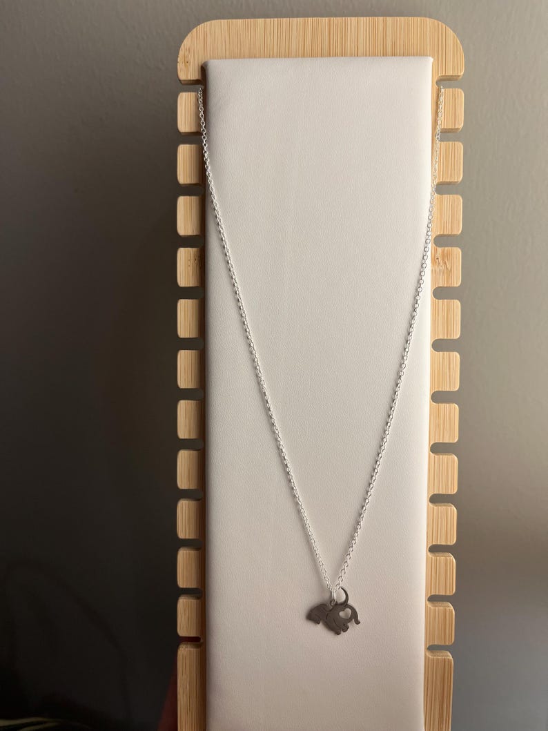 Puede incluir: Un collar de cadena de plata con un peque&ntilde;o colgante de elefante de plata. El collar se muestra en un soporte de madera.