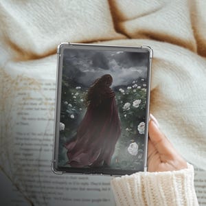 May include: A clear e-reader case with a fantasy art print. The image shows a person in a red cloak amidst white roses under a cloudy sky. The case is held by a person wearing a cream-coloured sweater. The case is designed to protect the e-reader.