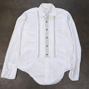 Pode incluir: Camisa branca de manga comprida com frente pregueada e fecho de botões pretos. A camisa apresenta painéis verticais decorativos com um padrão de pontos em ambos os lados das pregas. Os punhos são abotoados e a gola é pontiaguda.