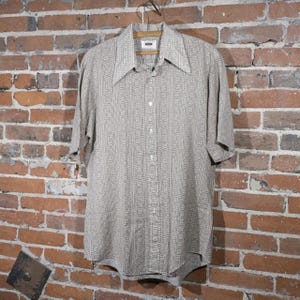 May include: A short-sleeved button-down shirt with a geometric pattern in shades of beige and off-white. The shirt has a wide collar and is hanging on a wooden hanger against a brick wall. The shirt is a vintage style.