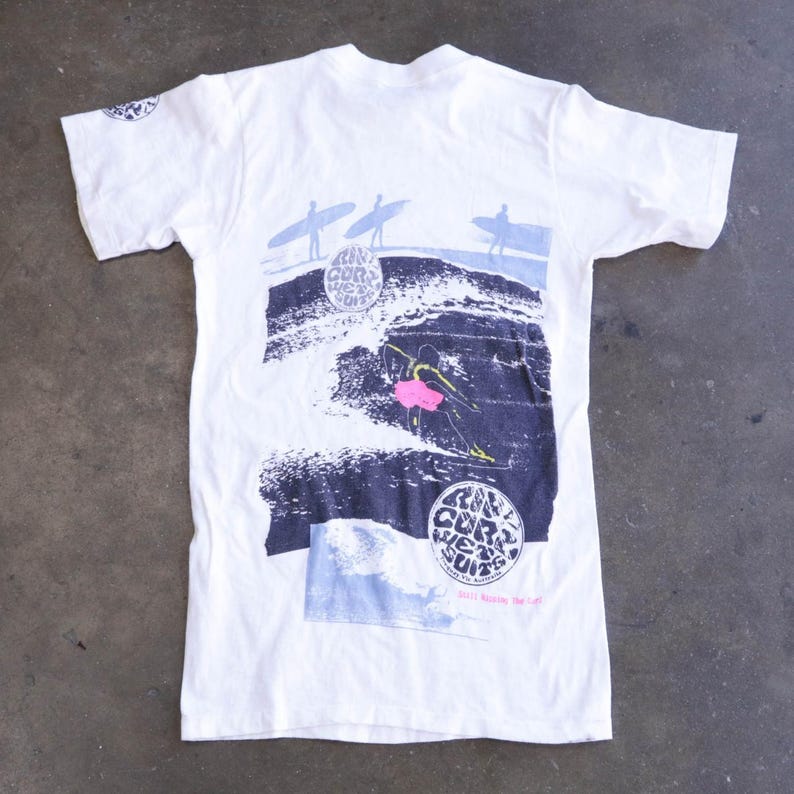 May include: White t-shirt featuring a surfing graphic. The design includes surfers on waves, a surfer in pink, and the text "Ain't Cor Wet Suits". The shirt has a crew neck and short sleeves.