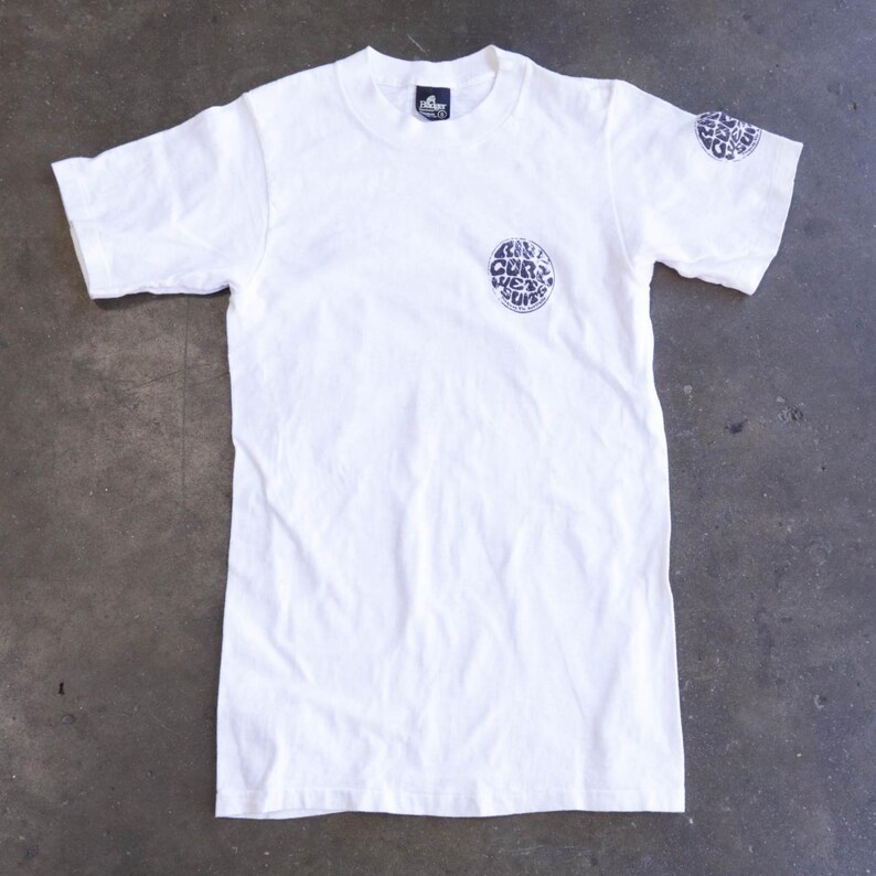 May include: White t-shirt with short sleeves. The shirt features a circular graphic on the chest and upper left sleeve with the text "RILL CORP WET SUITS" in a dark blue color. A small tag is visible at the neckline.