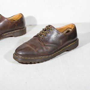 May include: A pair of dark brown leather oxford shoes with a rounded toe, lace-up closure, and yellow stitching along the thick, dark brown rubber sole. The interior lining is tan, and the shoes feature a worn, textured finish on the leather surface.