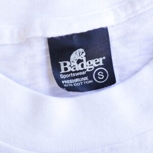 May include: Close-up of a white t-shirt with a black tag. The tag reads "Badger Sportswear" with a badger logo and "PRESHRUNK 100% COTTON" text. A small "S" is in a white circle.