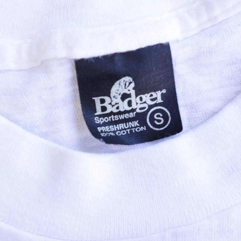 May include: Close-up of a white t-shirt with a black tag. The tag reads "Badger Sportswear" with a badger logo and "PRESHRUNK 100% COTTON" text. A small "S" is in a white circle.