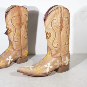 May include: A pair of tan leather cowboy boots featuring a pointed toe and a stacked heel. The shaft is decorated with intricate yellow embroidery and a circular G logo. The foot area is adorned with multiple cream and yellow cross-shaped patches.