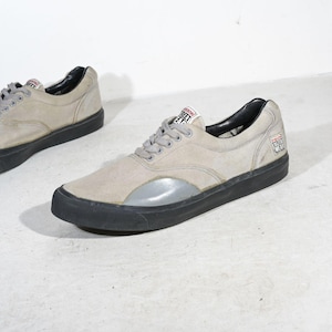 May include: A pair of low-top sneakers featuring light gray suede uppers, black rubber soles, and a glossy gray toe cap. The tongue and side heel display a white label with the red and black text Vision Street Wear. The interior is black.