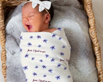 May include: A white swaddle blanket with a blue floral pattern and the text "Jesus, I trust in you" repeated throughout. A baby is wrapped in the blanket and is lying in a wicker basket.