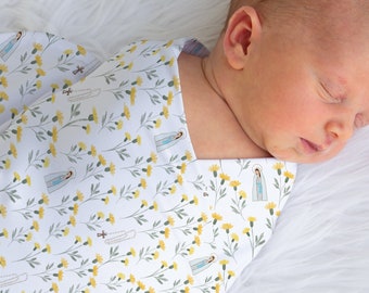 May include: A newborn baby swaddled in a white swaddle blanket with a yellow floral pattern. The baby is sleeping peacefully.