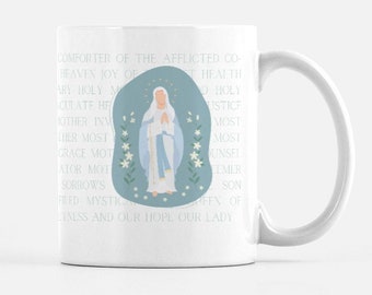 May include: White ceramic mug with a blue illustration of the Virgin Mary. The text around the image reads "COMFORTER OF THE AFFLICTED CO-HEAVEN JOY OF...LINESS AND OUR HOPE OUR LADY".