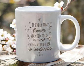 May include: White ceramic mug with a curved handle, featuring the quote "If every tiny flower wanted to be a rose, spring would lose its loveliness." The quote is in a handwritten style font. The mug is on a wooden surface.