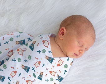May include: A newborn baby swaddled in a white muslin swaddle blanket with a colorful pattern of musical notes, crosses, and a woman playing a harp.